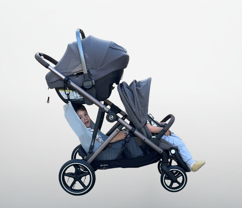 Gray and black triple stroller with a child inside the stroller sling hammock on a light gray background and a carseat on top. 