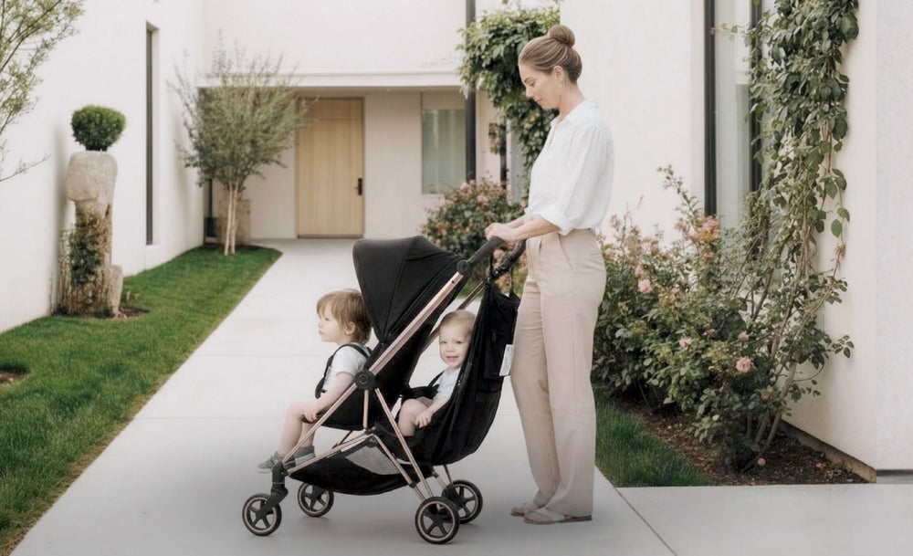 Woman pushing a stroller with two children on a sidewalk. One child is in the Stroller Sling hammock